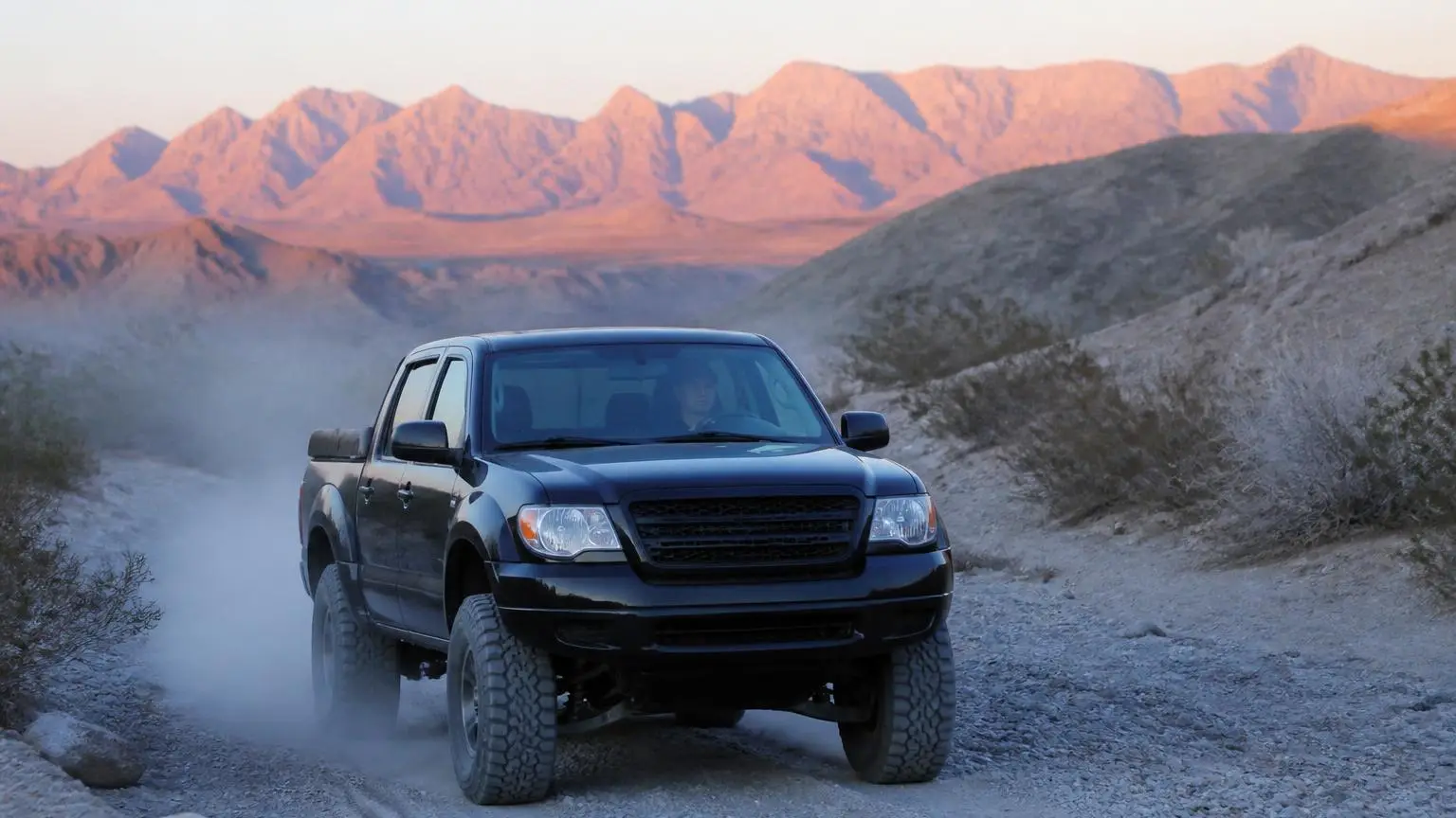 Black lifted mid-size pickup rolling through a desert wash at dawn on hybrid-tread tires